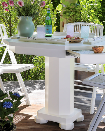Outdoor setting with a white game table and chairs on a wooden deck, surrounded by greenery.