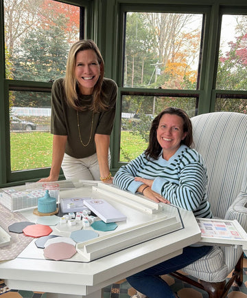 Two women sitting at a table with a large white tray containing various items, in front of a window with a view of trees and a fence.